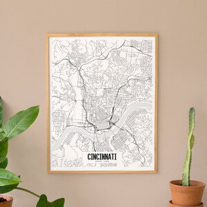 Puede incluir: Un mapa en blanco y negro enmarcado de Cincinnati, Ohio, con el nombre de la ciudad en la parte inferior. El mapa está en un marco de madera clara, exhibido sobre una superficie de madera con plantas en macetas, incluyendo una planta grande y cactus.