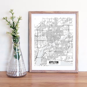 Puede incluir: Una impresión enmarcada en blanco y negro del mapa de Appleton, Wisconsin, EE. UU. El mapa detalla calles y vías fluviales, en un marco de madera clara. Un jarrón de cristal transparente con tallos florales blancos está a la izquierda.