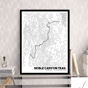 May include: A framed black and white topographic map print titled Noble Canyon Trail, 9.9 Miles, California, USA. The artwork features intricate black contour lines on a white background, encased in a simple black frame.
