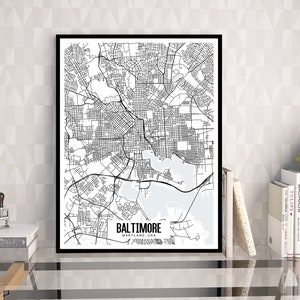 May include: A framed monochrome map print of Baltimore, Maryland, USA. The detailed city map shows streets, waterways, and the city's layout. The print is displayed on a glass table with books and a plant.