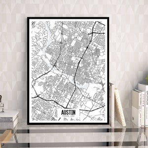 Puede incluir: Un mapa en blanco y negro enmarcado de Austin, Texas, EE. UU., exhibido sobre una mesa de vidrio. El mapa detalla las calles y vías fluviales de la ciudad. La palabra "AUSTIN" está impresa debajo. La imagen está sobre un fondo neutro.