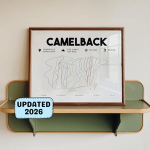 May include: A framed print of the Camelback ski resort map, with the resort name in bold black letters. The map shows ski runs in red and blue lines. The frame is brown, and the print is displayed on a green and wood-toned shelf. The text "UPDATED 2026" is on a blue sticker.