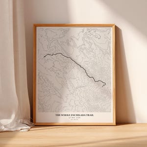 May include: Framed print of a topographic map of The Whole Enchilada Trail in Utah, USA. The map is in black and white, with a black line tracing the trail. The frame is a light wood color.