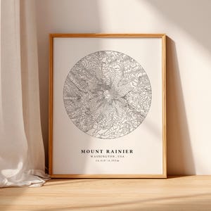 Op de afbeelding: Een ingelijste print met een cirkelvormige topografische kaart van Mount Rainier, Washington, VS. De kaart gebruikt zwarte contourlijnen op een crèmekleurige achtergrond. De tekst "MOUNT RAINIER WASHINGTON, USA 14,410 ft / 4,392m" staat eronder. De lijst is lichtbruin.