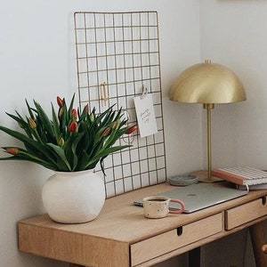 Peut inclure: Un bureau en bois clair avec deux tiroirs et une lampe dorée. Un ordinateur portable, une tasse et une pile de livres sont sur le bureau. Une chaise en osier avec un siège tissé est devant le bureau. Un panier en osier avec des anses en cuir est sur le sol. Un vase blanc avec des tulipes rouges est sur le bureau. Une grille en fil de fer doré avec des cartes est sur le mur. Un dessin au trait noir et blanc encadré de deux visages est sur le mur.