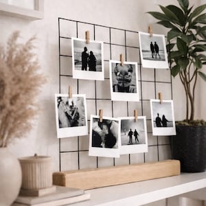 Può includere: Espositore fotografico a griglia metallica nera con diverse foto in bianco e nero fissate con piccole mollette di legno. L'espositore poggia su una base di legno. Una pianta in vaso e oggetti decorativi su una mensola.