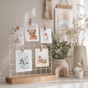 Può includere: Un espositore fotografico a filo bianco con base in legno contiene sei piccole foto fissate con mollette di legno. Le foto presentano immagini di un orsacchiotto, fiori e stampe botaniche. Una stampa artistica arcobaleno e altri oggetti decorativi sono sullo sfondo.