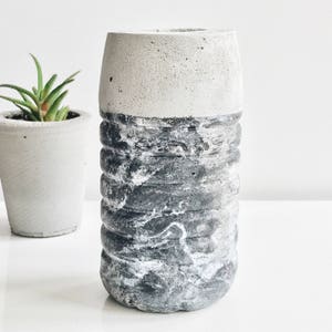 Op de afbeelding: Een grijze en witte betonnen vaas met een getextureerd, marmerachtig ontwerp. De vaas is cilindrisch en heeft een brede opening aan de bovenkant.