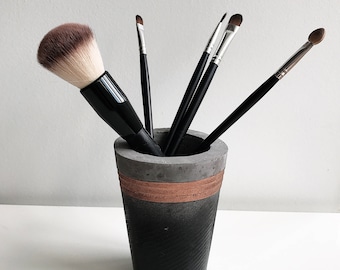 May include: A gray concrete makeup brush holder with a copper band around the middle. Five black makeup brushes are in the holder.