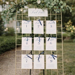 Puede incluir: Una rejilla metálica dorada que muestra las asignaciones de mesa para una boda. Cada tarjeta está numerada y atada con una cinta azul marino. La tarjeta superior dice "Find Table". La vegetación y las flores blancas adornan la parte superior.
