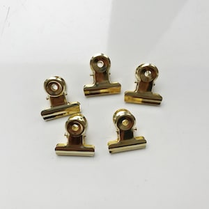 Puede incluir: Cinco clips de carpeta dorados sobre una superficie blanca. Los clips son todos del mismo tamaño y forma, con una cabeza redonda y dos brazos que se pueden usar para sujetar papeles juntos.