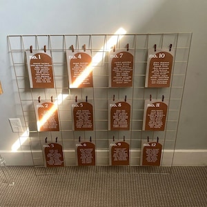 Puede incluir: Una rejilla de alambre dorada con 12 tarjetas, cada una con un número y una lista de nombres. Las tarjetas son marrones con texto blanco y tienen una forma de arco redondeado en la parte superior. Las tarjetas están etiquetadas con los números del 1 al 12.