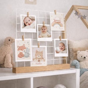 Può includere: Un espositore fotografico a filo bianco con clip in legno che contengono foto di bambini e biglietti illustrati. Le carte presentano animali e arcobaleni. Una mensola in legno sottostante contiene giocattoli e oggetti decorativi, creando un tema di cameretta.