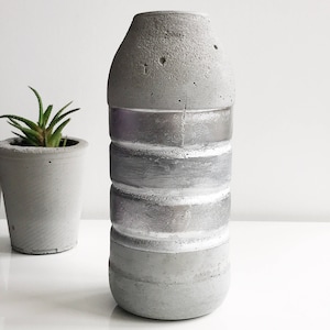 May include: A grey concrete vase with a silver metallic finish. The vase has a cylindrical shape with a rounded top and bottom. It is decorated with horizontal bands of silver paint.
