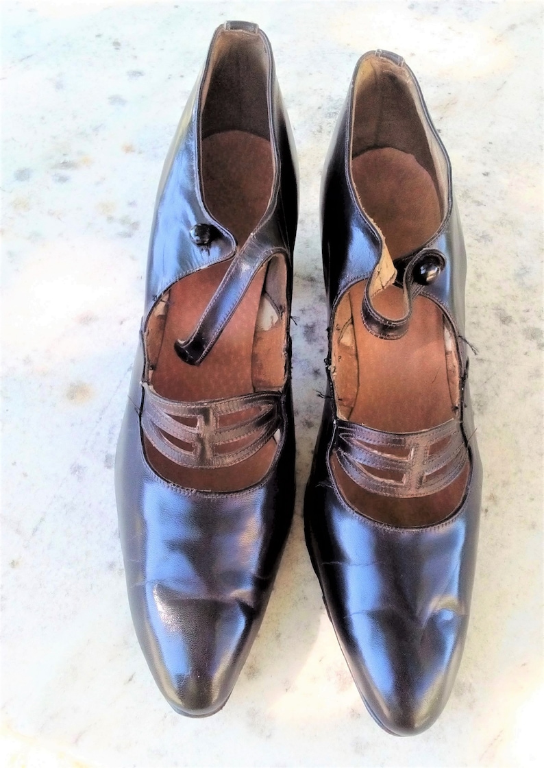 May include: A pair of black leather shoes with a pointed toe and a strap across the instep. The shoes have a decorative cut-out design on the strap.
