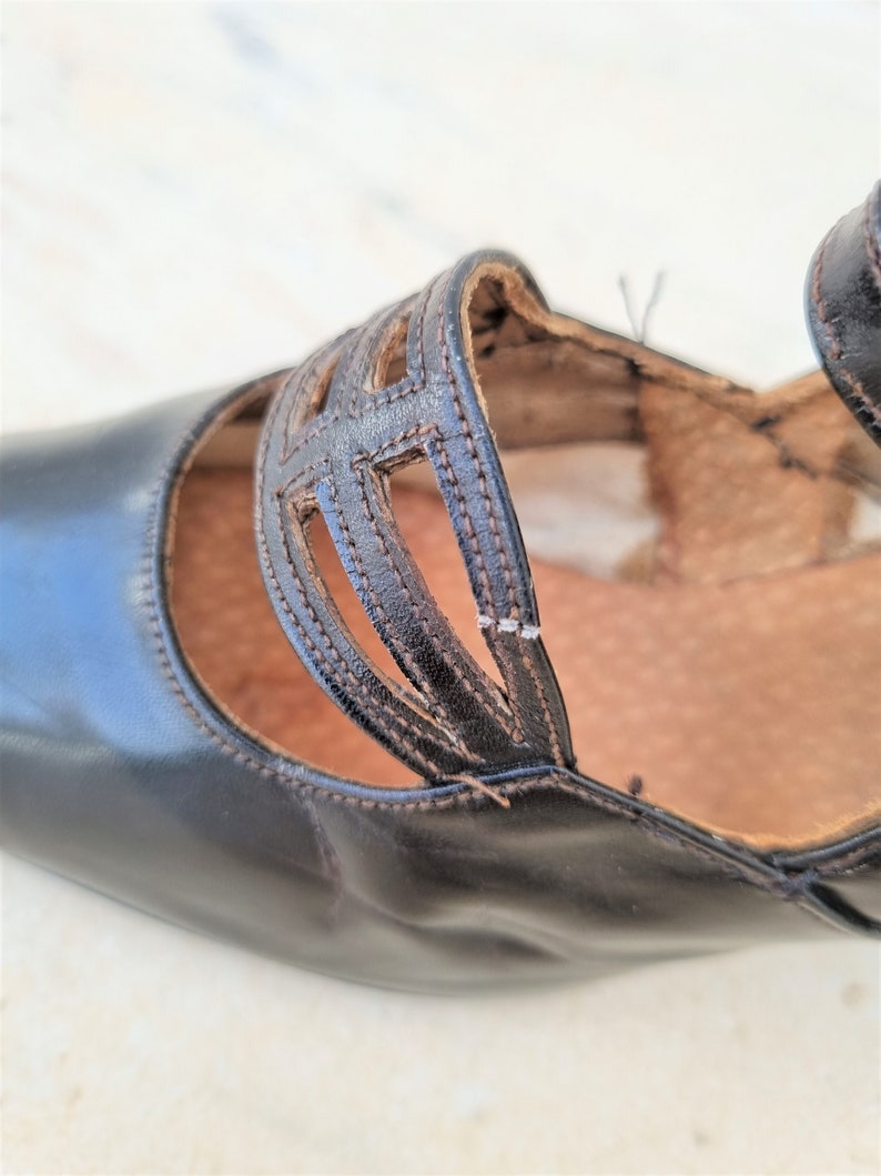 May include: Close-up of a black leather shoe with a cut-out design on the strap. The shoe has a pointed toe and a low heel.