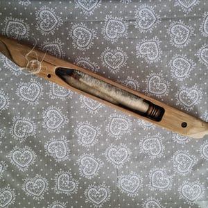 May include: A wooden bobbin with a white thread wound around it. The bobbin is made of light brown wood and has a small metal screw on the side. The bobbin is lying on a gray fabric with a white heart pattern.