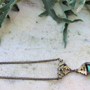 Peut inclure: Un collier pendentif vintage au design d&eacute;taill&eacute;. Le collier pr&eacute;sente une cha&icirc;ne d&eacute;licate et un pendentif avec une pierre vert &eacute;meraude. Le pendentif est encadr&eacute; par un sertissage or antique orn&eacute;. L'arri&egrave;re-plan comprend des feuilles vertes et une surface de couleur claire.