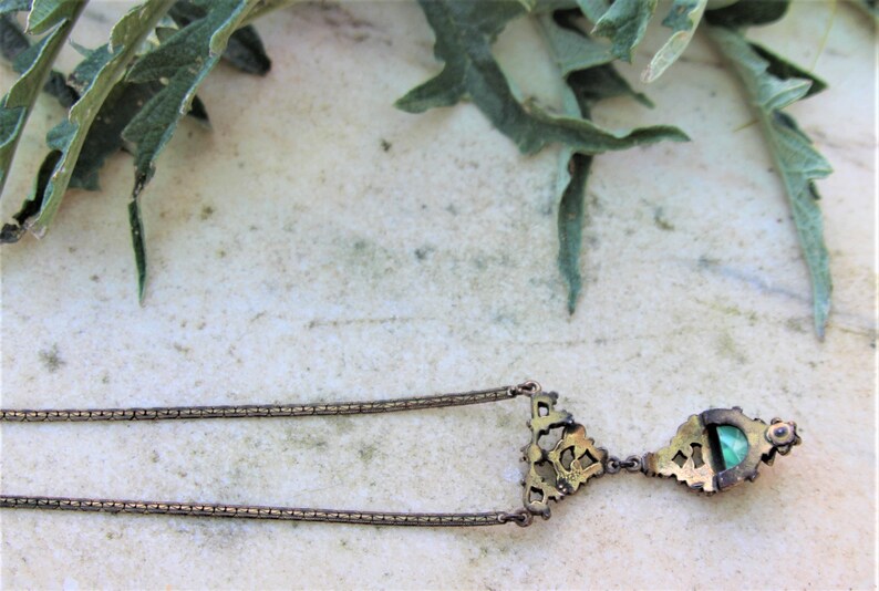Peut inclure: Un collier pendentif vintage au design d&eacute;taill&eacute;. Le collier pr&eacute;sente une cha&icirc;ne d&eacute;licate et un pendentif avec une pierre vert &eacute;meraude. Le pendentif est encadr&eacute; par un sertissage or antique orn&eacute;. L'arri&egrave;re-plan comprend des feuilles vertes et une surface de couleur claire.
