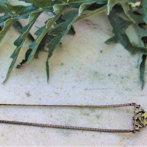 Peut inclure: Un collier dor&eacute; antique avec une cha&icirc;ne rectangulaire et un pendentif d&eacute;coratif. Le pendentif pr&eacute;sente un design orn&eacute; avec une petite pierre pr&eacute;cieuse verte carr&eacute;e. Des feuilles vertes sont en arri&egrave;re-plan.