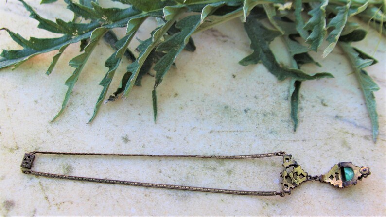 Peut inclure: Un collier dor&eacute; antique avec une cha&icirc;ne rectangulaire et un pendentif d&eacute;coratif. Le pendentif pr&eacute;sente un design orn&eacute; avec une petite pierre pr&eacute;cieuse verte carr&eacute;e. Des feuilles vertes sont en arri&egrave;re-plan.