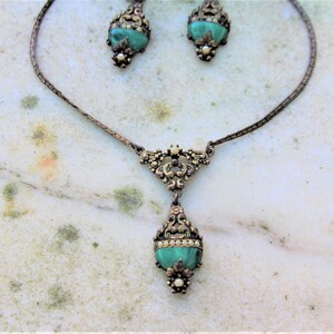 Peut inclure: Un ensemble de bijoux anciens comprenant un collier et des boucles d'oreilles assorties. L'ensemble pr&eacute;sente un design ornemental en m&eacute;tal de couleur bronze. Chaque pi&egrave;ce incorpore une pierre turquoise ovale. Le collier a un pendentif en forme de goutte.