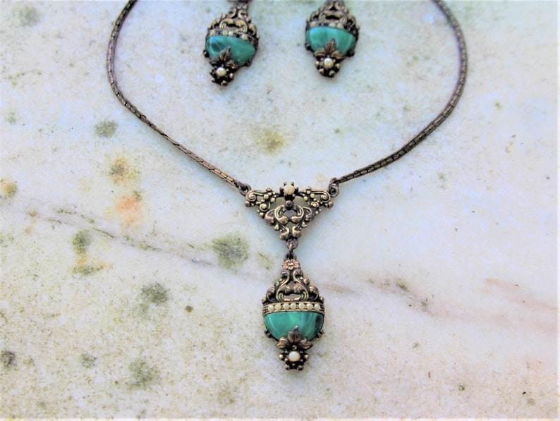 Peut inclure: Un ensemble de bijoux anciens comprenant un collier et des boucles d'oreilles assorties. L'ensemble pr&eacute;sente un design ornemental en m&eacute;tal de couleur bronze. Chaque pi&egrave;ce incorpore une pierre turquoise ovale. Le collier a un pendentif en forme de goutte.