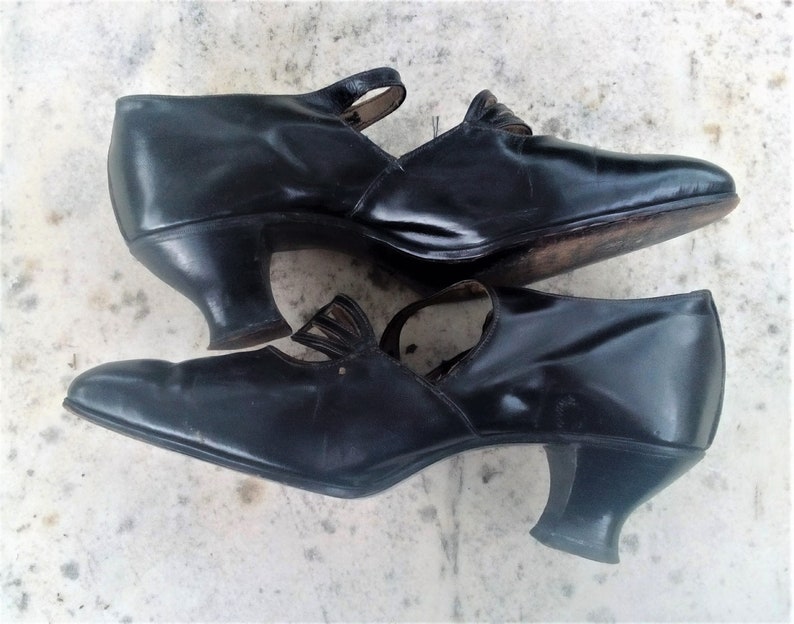 May include: A pair of black leather heeled shoes with a strap and buckle detail. The shoes have a pointed toe and a low heel.