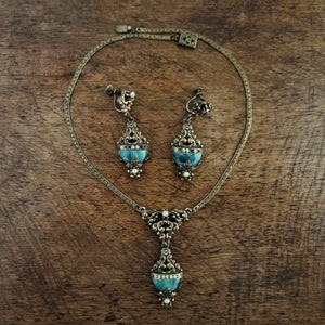 Peut inclure: Un ensemble de bijoux anciens comprenant un collier et des boucles d'oreilles &agrave; clip. L'ensemble est en m&eacute;tal dor&eacute; avec des d&eacute;tails de filigrane orn&eacute;s. Chaque pi&egrave;ce pr&eacute;sente une pierre de couleur turquoise et de petits accents blancs. Les bijoux sont pr&eacute;sent&eacute;s sur une surface en bois.