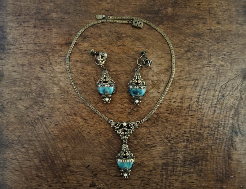 Peut inclure: Un ensemble de bijoux anciens comprenant un collier et des boucles d'oreilles &agrave; clip. L'ensemble est en m&eacute;tal dor&eacute; avec des d&eacute;tails de filigrane orn&eacute;s. Chaque pi&egrave;ce pr&eacute;sente une pierre de couleur turquoise et de petits accents blancs. Les bijoux sont pr&eacute;sent&eacute;s sur une surface en bois.