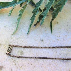 Peut inclure: Un collier dor&eacute; antique avec un fermoir rectangulaire orn&eacute; d'un motif floral. La cha&icirc;ne est compos&eacute;e de petits maillons interconnect&eacute;s. Des feuilles vertes sont visibles en arri&egrave;re-plan, ajoutant un &eacute;l&eacute;ment naturel &agrave; la composition.