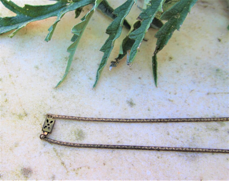Peut inclure: Un collier dor&eacute; antique avec un fermoir rectangulaire orn&eacute; d'un motif floral. La cha&icirc;ne est compos&eacute;e de petits maillons interconnect&eacute;s. Des feuilles vertes sont visibles en arri&egrave;re-plan, ajoutant un &eacute;l&eacute;ment naturel &agrave; la composition.