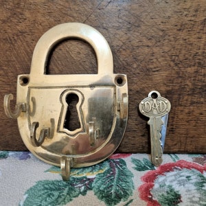 Op de afbeelding: Een messing sleutelhanger in de vorm van een hangslot met een sleutelgat en vier haken. De sleutelhanger ligt op een houten oppervlak. Een kleine messing sleutel met het woord "DAD" erop gegraveerd ligt naast de sleutelhanger.