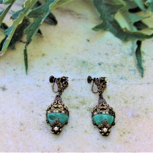 Peut inclure: Une paire de boucles d'oreilles vintage &agrave; clip. Chaque boucle d'oreille pr&eacute;sente une pierre verte en forme de goutte d'eau sertie dans un cadre filigran&eacute; dor&eacute; orn&eacute;. De petits accents en forme de perles sont visibles au bas de chaque boucle d'oreille.