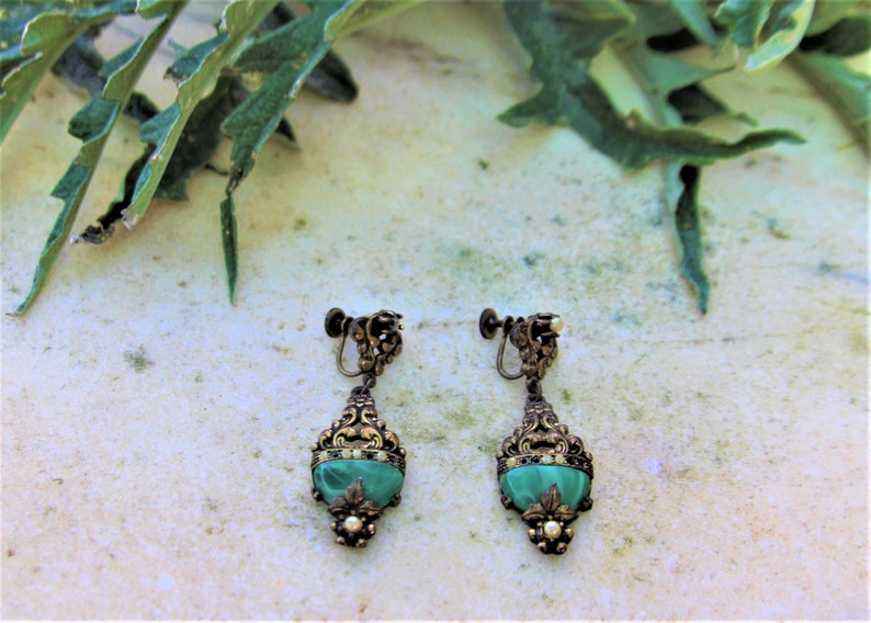 Peut inclure: Une paire de boucles d'oreilles vintage &agrave; clip. Chaque boucle d'oreille pr&eacute;sente une pierre verte en forme de goutte d'eau sertie dans un cadre filigran&eacute; dor&eacute; orn&eacute;. De petits accents en forme de perles sont visibles au bas de chaque boucle d'oreille.