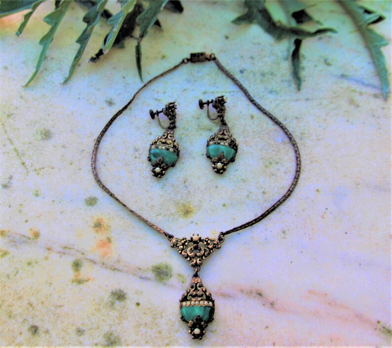 Peut inclure: Ensemble de bijoux vintage comprenant un collier et des boucles d'oreilles assorties. L'ensemble pr&eacute;sente une finition en m&eacute;tal fonc&eacute; avec des d&eacute;tails de filigrane orn&eacute;s. Chaque pi&egrave;ce met en valeur une pierre de couleur turquoise, orn&eacute;e de petites perles blanches et de strass transparents.