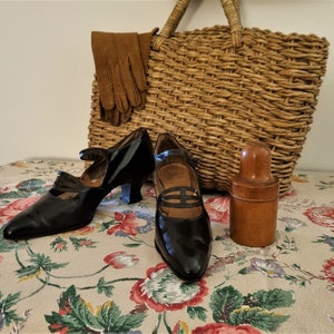May include: Two pairs of black leather shoes with a pointed toe and a small heel. The shoes have a strap across the top and a buckle closure. The shoes are sitting on a floral tablecloth with a wicker basket and a wooden bottle in the background.