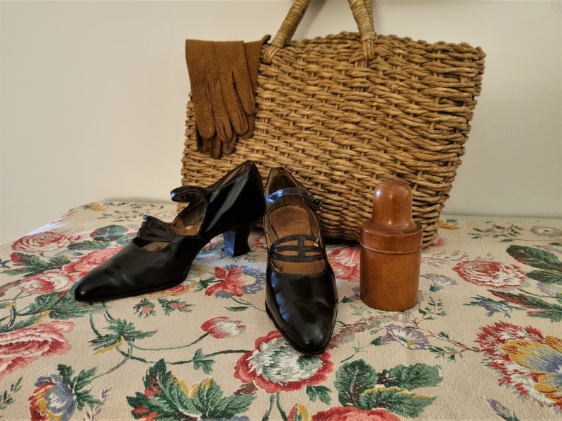 May include: Two pairs of black leather shoes with a pointed toe and a small heel. The shoes have a strap across the top and a buckle closure. The shoes are sitting on a floral tablecloth with a wicker basket and a wooden bottle in the background.