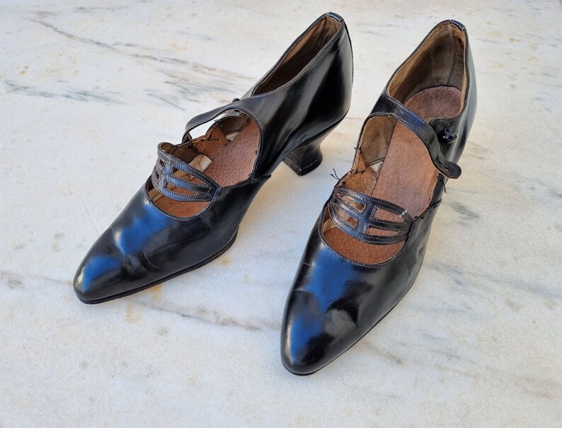 May include: A pair of black leather heeled shoes with a strap across the instep and a decorative cut-out design.