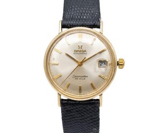 Può includere: Un orologio da polso automatico Omega Seamaster De Ville con cassa rotonda color oro, quadrante argentato con indici dorati, datario a ore tre e cinturino in pelle nera testurizzata con motivo a pelle di lucertola.