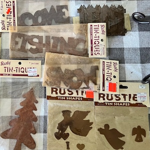 May include: A collection of Rusty Tin-Tiques metal cutouts in individual plastic packaging. The set includes word cutouts like HALLOWEEN, GONE FISHING, and SNOW, along with shapes of a picket fence, a pine tree, and various angel silhouettes.