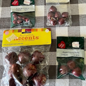 May include: Several packages of craft supplies are displayed on a checkered surface. The packages contain miniature red apples and cherries, labeled as "Apple-Micro Burg." and "Apple/Cherry Burg." The packages are from Lara's Crafts and Craft Connection.