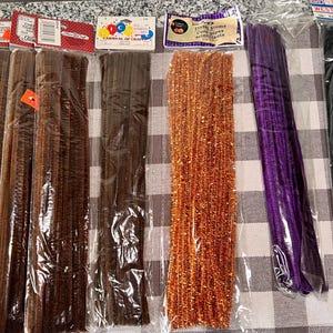 May include: Assortment of colorful pipe cleaners in brown, orange, purple, and black. The pipe cleaners are packaged in clear plastic bags with labels.