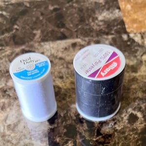 May include: Two spools of sewing thread. One spool is white with a blue label reading Dual Duty XP All Purpose 250 YDS. The other spool is black with a white and red label reading Allary 100% Polyester 182 M.