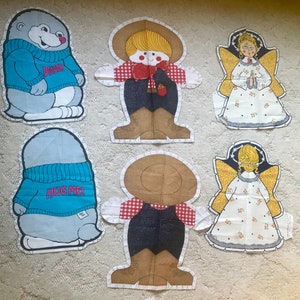 May include: A collection of fabric appliques featuring whimsical designs. Includes a mole wearing a blue sweater with "DIGGER" and "HUG ME!" text, a cowboy, and two angels with yellow wings. The appliques are outlined with a white, scalloped edge.