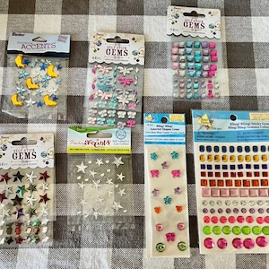 May include: A collection of self-stick gems in various shapes and colors, including stars, moons, butterflies, and squares. Packages include the text "Self Stick Gems" and "Finishing Accents".
