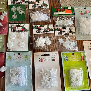 May include: A collection of various packages of white snowflake-shaped Christmas ornaments. Each package contains multiple small plastic snowflake decorations, some with hanging strings, displayed on branded cardboard backing cards.