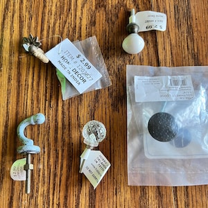 May include: A collection of decorative cabinet knobs and pulls in various styles. Includes a clear glass globe knob, a white ceramic knob, a black textured knob, and a light blue hook-shaped pull. Some items have price tags.