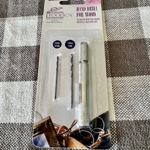 May include: A hand drill for wood, packaged in a clear plastic blister pack. The package includes a silver-colored hand drill with a knurled handle and two drill bits. The text on the package reads "CRAFTER'S TOOLBOX" and "HAND DRILL FOR WOOD".