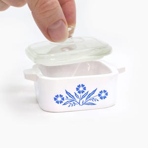 May include: A small, white casserole dish with a clear glass lid. The dish features a blue floral design. The lid is being lifted by a hand. The dish is square with small handles on each side.